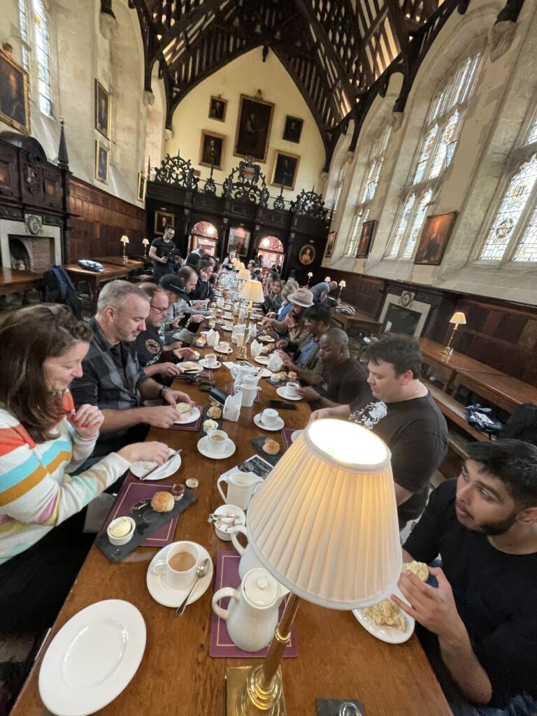 The dining hall of Exeter College, Oxford, where attendees are tucking into scones, clotted cream and jam. They sit at long wooden tables, with high vaulted ceiling above them.